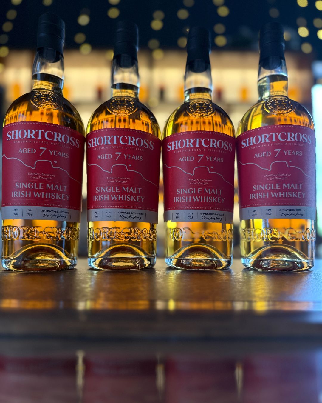 Four bottles of Shortcross 7-year-old Single Malt Irish Whiskey on a wooden surface with a blurred bar background.