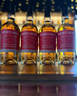 Four bottles of Shortcross 7-year-old Single Malt Irish Whiskey on a wooden surface with a blurred bar background.